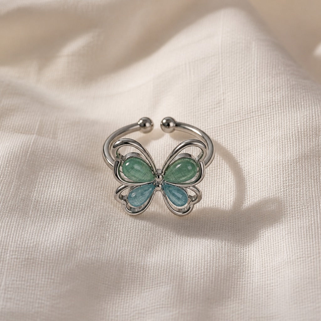 Open band butterfly ring with glossy blue and green teardrop stones, on a cream fabric