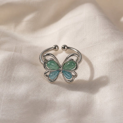 Open band butterfly ring with glossy blue and green teardrop stones, on a cream fabric