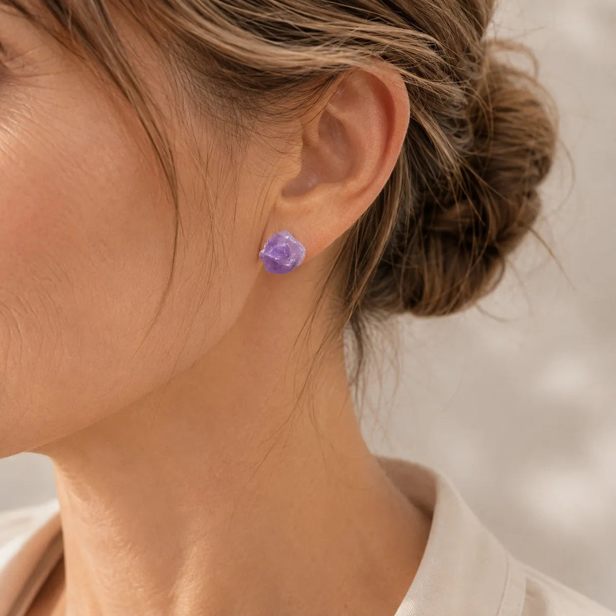 Close-up of a woman's ear wearing a small raw-cut purple amethyst stud earring with her hair in an updo