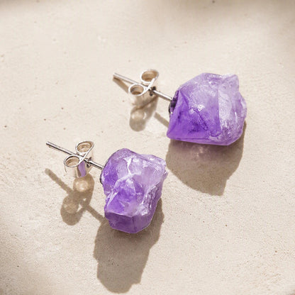 Pair of raw-cut purple amethyst stud earrings with post backs on a pale stone surface